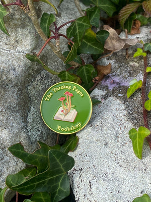 The Turning Pages Enamel pin sits on a rock outside